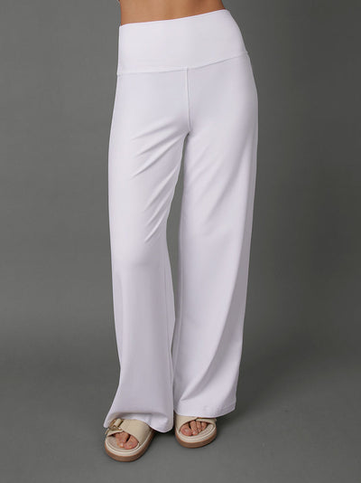 A model wearing a white two-piece activewear set consisting of a sports bra and high-waisted, wide-leg yoga pants, paired with beige slide sandals, set against a solid gray studio background.