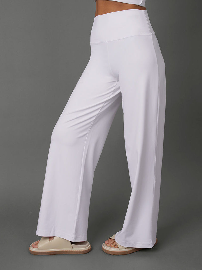 White high-waisted wide-leg pants worn by a model, paired with a matching white crop top and beige slide sandals, set against a solid gray studio background.