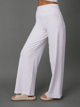 White high-waisted wide-leg pants worn by a model, paired with a matching white crop top and beige slide sandals, set against a solid gray studio background.
