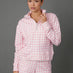 A woman modeling a pink and white gingham check two-piece loungewear set, consisting of a long-sleeve quarter-zip pullover and matching shorts, against a solid gray studio background.