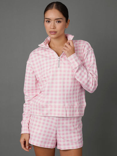 A woman modeling a pink and white gingham check two-piece loungewear set, consisting of a long-sleeve quarter-zip pullover and matching shorts, against a solid gray studio background.