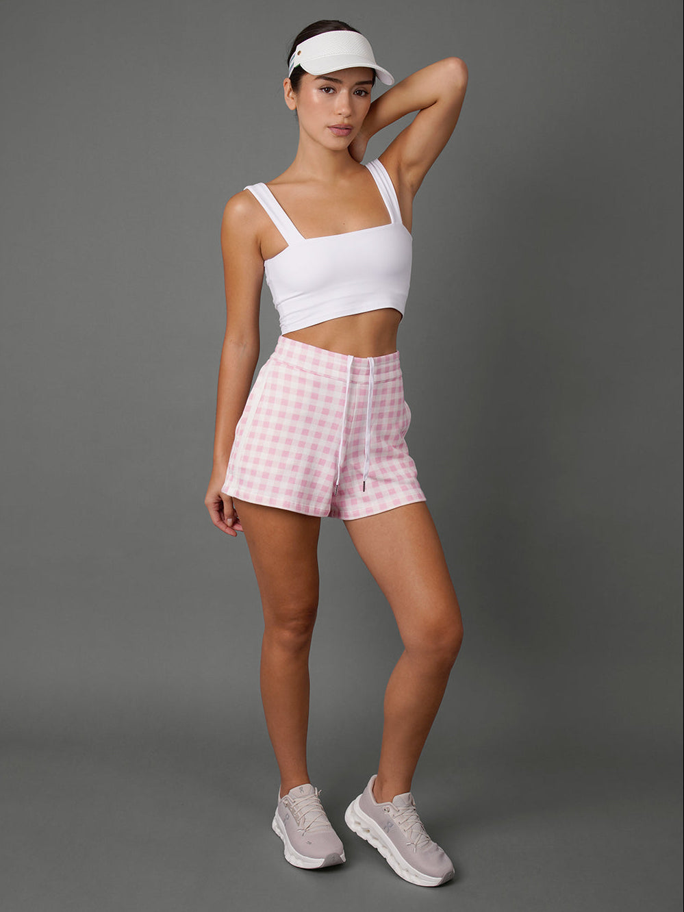 Model wears a white sports bra, a  Pink Gingham french terry sweat short, and a white visor against a grey background.