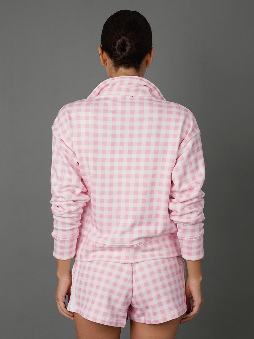 A woman viewed from behind wearing a pink and white gingham check long-sleeve pajama top and matching shorts against a solid gray studio background.