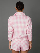 A woman viewed from behind wearing a pink and white gingham check long-sleeve pajama top and matching shorts against a solid gray studio background.