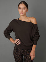 A woman modeling a dark brown off-the-shoulder long-sleeve sweatshirt with a thin black spaghetti strap detail, paired with leopard print leggings, against a solid gray studio background.