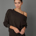 A woman modeling a dark brown off-the-shoulder long-sleeve sweatshirt with a thin black underband and black leopard-print leggings, set against a solid gray studio background.