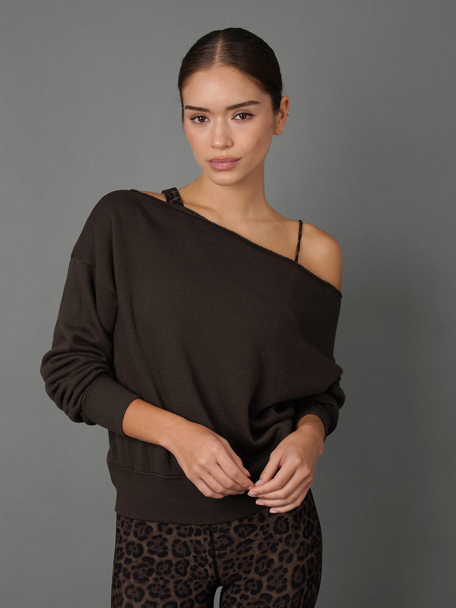 A woman modeling a dark brown off-the-shoulder long-sleeve sweatshirt with a thin black underband and black leopard-print leggings, set against a solid gray studio background.