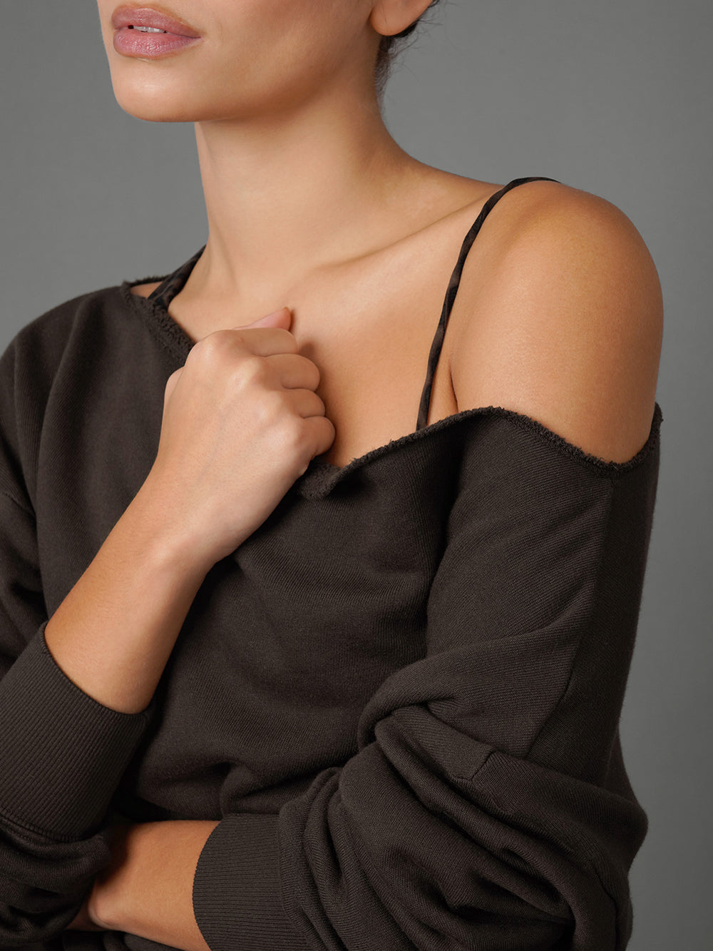 A close-up of a model wearing a dark brown off-the-shoulder knit top with thin spaghetti straps over a black camisole, set against a solid gray studio background.