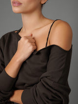 A close-up of a model wearing a dark brown off-the-shoulder knit top with thin spaghetti straps over a black camisole, set against a solid gray studio background.