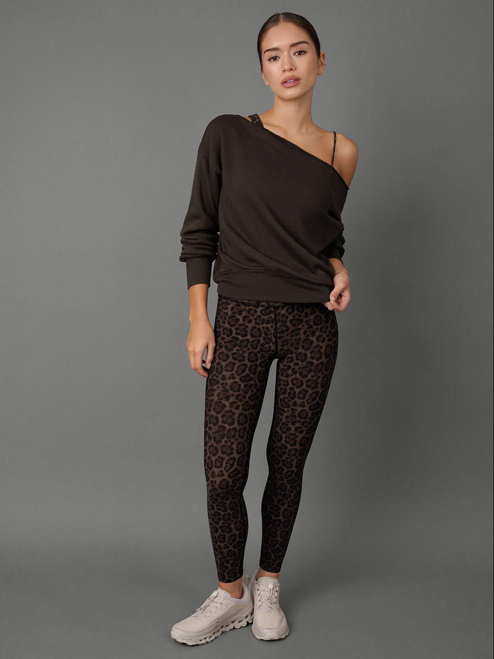 A woman modeling a dark brown long-sleeve asymmetrical sweater and brown leopard-print leggings with white sneakers, standing against a solid gray studio background.