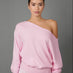 A woman modeling a light pink one-shoulder sweatshirt with a relaxed fit and ribbed cuffs and hem, paired with pink and white gingham pants, against a solid gray studio background.