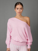 A woman modeling a light pink one-shoulder sweatshirt with a relaxed fit and ribbed cuffs and hem, paired with pink and white gingham pants, against a solid gray studio background.