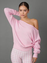 A woman modeling a light pink off-the-shoulder long-sleeve sweatshirt paired with pink and white gingham leggings, set against a solid dark gray studio background.