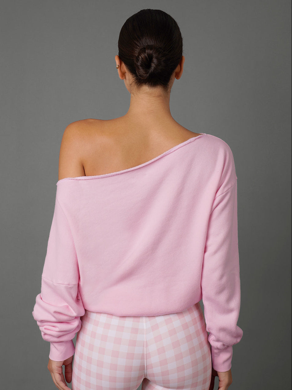 A woman shown from the back wearing a light pink off-the-shoulder long-sleeve top and pink-and-white gingham pants against a solid gray studio background.