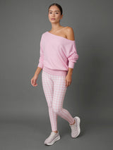 A woman modeling a light pink off-the-shoulder long-sleeve top and pink-and-white gingham leggings with white sneakers, standing against a solid gray studio background.