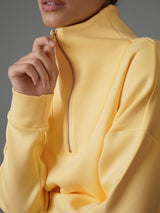 Closeup of a model holding the zipper on an interlock half zip sweatshirt in yellow against a grey backdrop. 