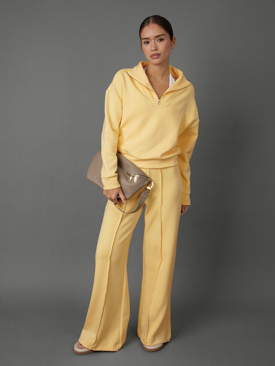 Model wears an interlock half zip sweatshirt and wide leg interlock pants in yellow against a grey backdrop. 