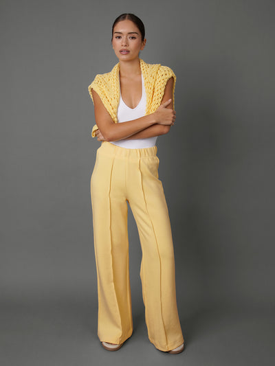 Model wears yellow interlock pants with a white tank top and a yellow sweater on her shoulders against a grey background.