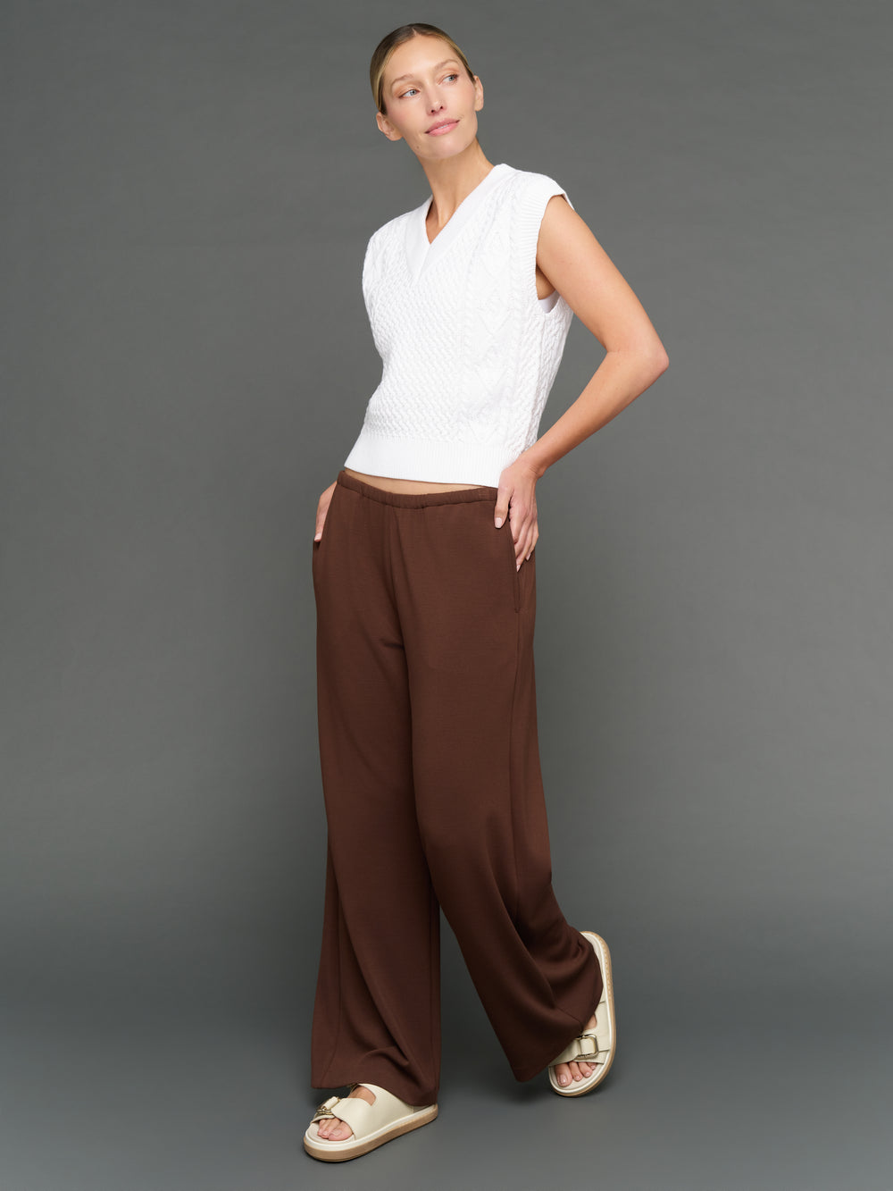 Woman wearing a white top and brown pants against a gray background