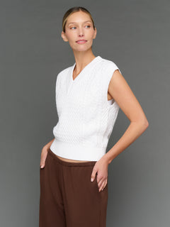 Woman wearing a white sleeveless knitted top and brown pants against a gray background