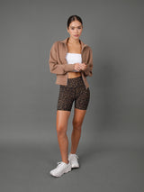 A woman stands against a solid gray studio background wearing a cropped tan zip-up hoodie, leopard-print biker shorts, a white tube top, and white sneakers.