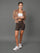 A woman stands against a solid gray studio background wearing a cropped tan zip-up hoodie, leopard-print biker shorts, a white tube top, and white sneakers.