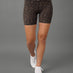 A model from the waist down wearing high-waisted leopard print athletic shorts, a white crop top, and white sneakers against a solid gray studio background.