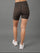 Rear view of a model wearing leopard-print biker shorts, a white crop top, and white sneakers against a solid gray studio background.