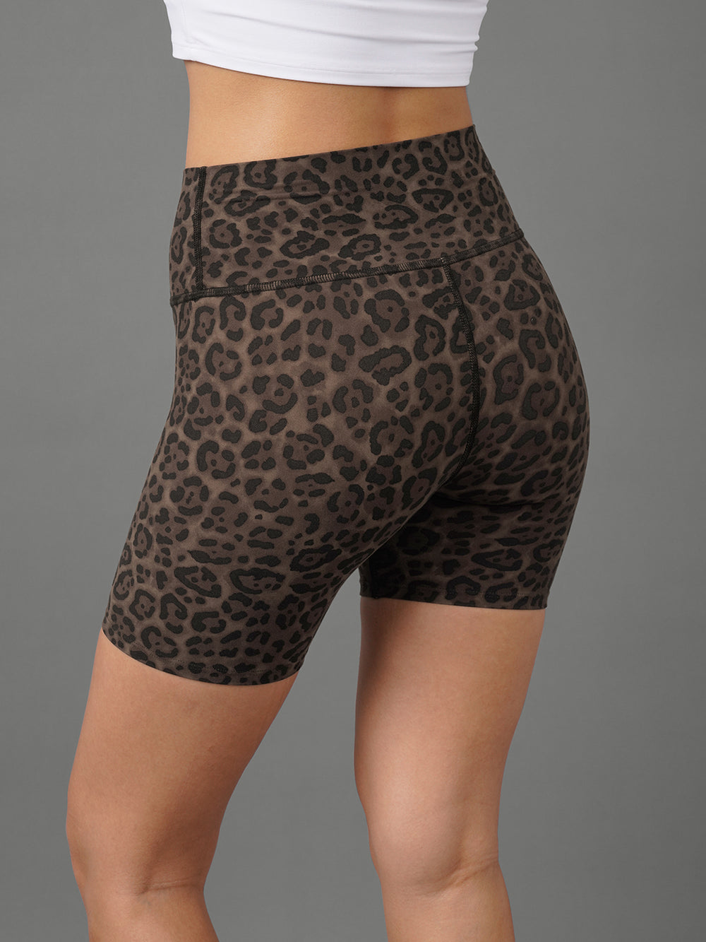 A close-up of a model wearing high-waisted leopard print biker shorts in brown and black, paired with a white crop top, shown from a rear three-quarter angle against a solid gray background.