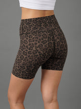A close-up of a model wearing high-waisted leopard print biker shorts in brown and black, paired with a white crop top, shown from a rear three-quarter angle against a solid gray background.