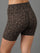 A close-up of a model wearing high-waisted leopard print biker shorts in brown and black, paired with a white crop top, shown from a rear three-quarter angle against a solid gray background.