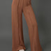 A model wearing high-waisted, wide-leg brown pants with a gathered elastic waistband, paired with white slide sandals, set against a solid dark gray studio background.