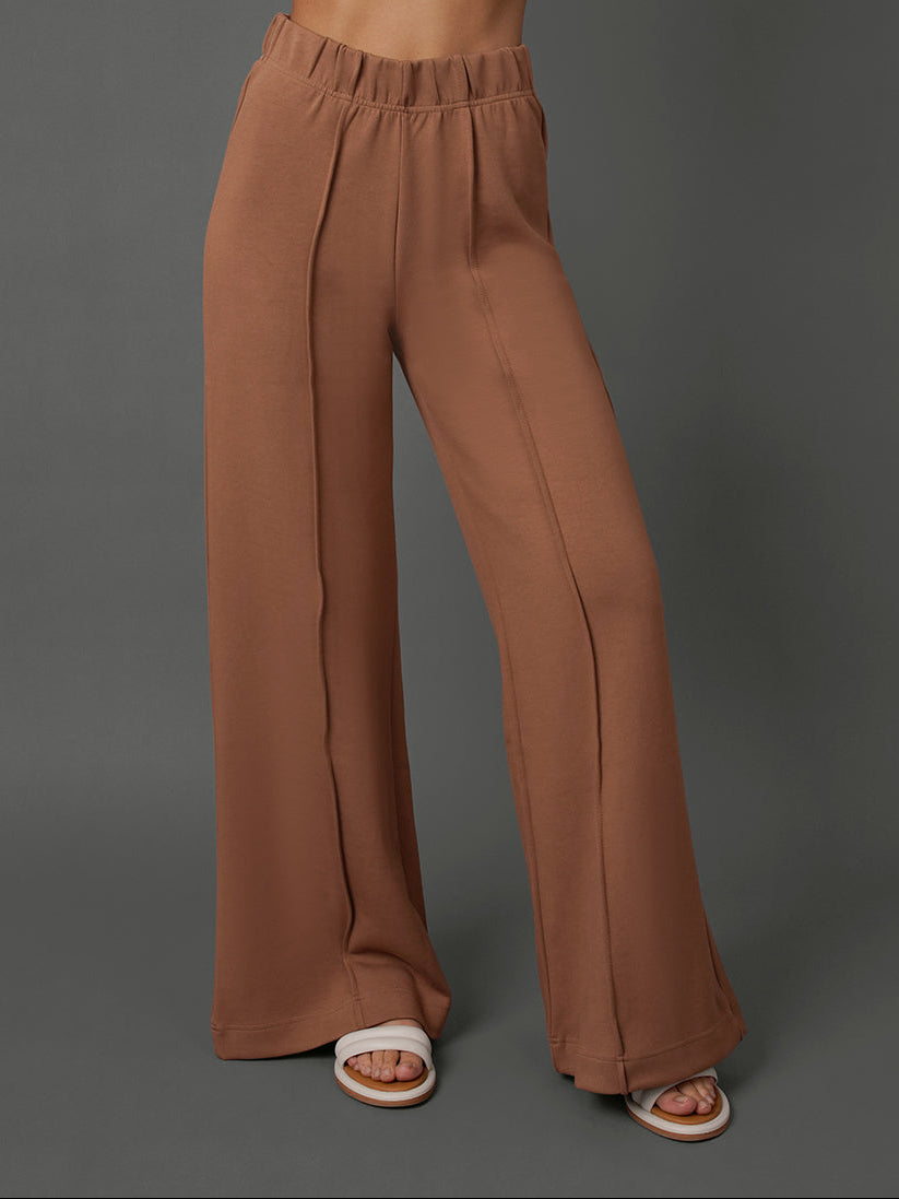 A model wearing high-waisted, wide-leg brown pants with a gathered elastic waistband, paired with white slide sandals, set against a solid dark gray studio background.