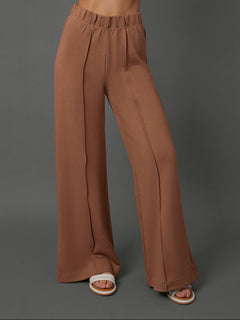 A model wearing high-waisted, wide-leg brown pants with a gathered elastic waistband, paired with white slide sandals, set against a solid dark gray studio background.