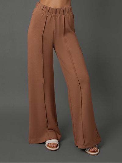 A model wearing high-waisted, wide-leg brown pants with a gathered elastic waistband, paired with white slide sandals, set against a solid dark gray studio background.