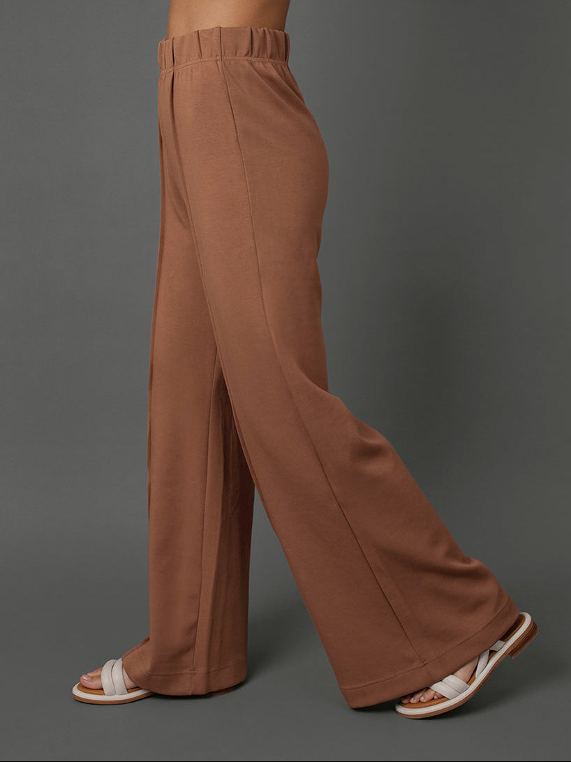 A model wearing high-waisted, wide-leg brown pants, shown from the side against a solid gray studio background.