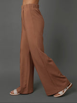 A model wearing high-waisted, wide-leg brown pants, shown from the side against a solid gray studio background.