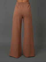 A model shown from the back wearing high-waisted, wide-leg pants in a warm terracotta brown color, against a solid dark gray studio background.