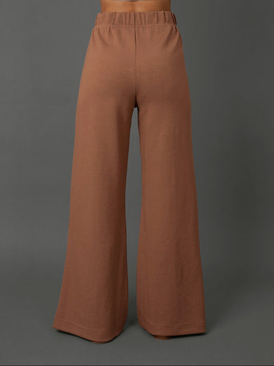 A model shown from the back wearing high-waisted, wide-leg pants in a warm terracotta brown color, against a solid dark gray studio background.