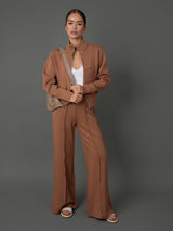 A woman modeling a matching two-piece brown suit consisting of a cropped zip-front jacket and wide-leg trousers, styled with a white top, a beige shoulder bag, and white slide sandals against a solid dark gray studio background.