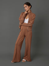 A model wearing a matching two-piece brown set consisting of a cropped zip-front jacket and wide-leg trousers, styled with a white tank top and white slide sandals against a solid gray studio background.