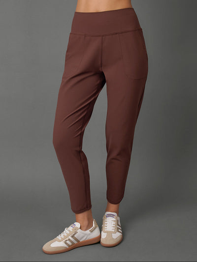 A model wearing a matching brown activewear set consisting of a cropped tank top and high-waisted leggings, paired with white sneakers, against a solid gray studio background.