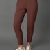 A model wearing a matching brown activewear set consisting of a cropped tank top and high-waisted leggings, paired with beige and white sneakers, against a solid gray studio background.