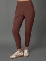 A model wearing a matching brown activewear set consisting of a cropped tank top and high-waisted leggings, paired with beige and white sneakers, against a solid gray studio background.