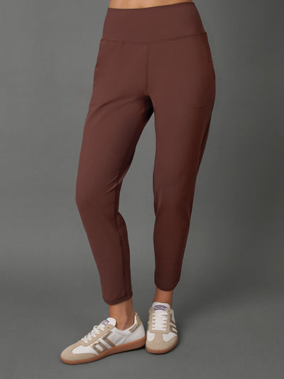 A model wearing a matching brown activewear set consisting of a cropped tank top and high-waisted leggings, paired with beige and white sneakers, against a solid gray studio background.