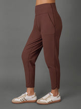 A model wearing high-waisted, ankle-length brown leggings with side pockets, paired with a matching brown sports bra and white sneakers, set against a solid gray studio background.