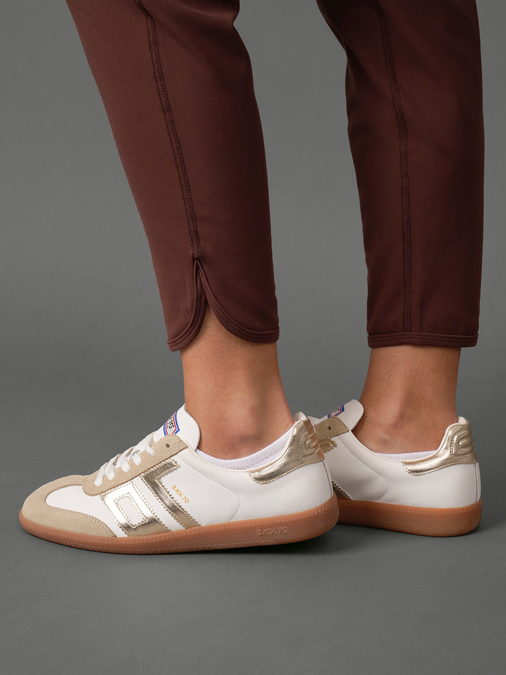 White leather sneakers with metallic gold accents and gum soles, shown on a model wearing brown trousers against a gray studio background.