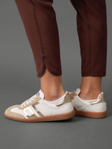 White leather sneakers with metallic gold accents and gum soles, shown on a model wearing brown trousers against a gray studio background.