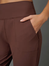 Close-up of a person wearing brown high-waisted leggings with a side pocket, showing the waistband, fabric texture, and hand in the pocket against a dark gray background.