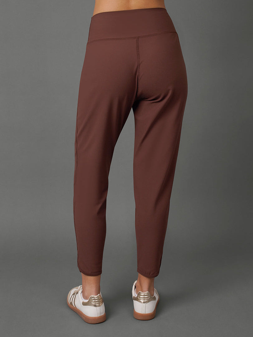 A model shown from the back wearing a matching brown activewear set, featuring high-waisted leggings and a cropped sports bra, paired with white sneakers, against a solid gray studio background.